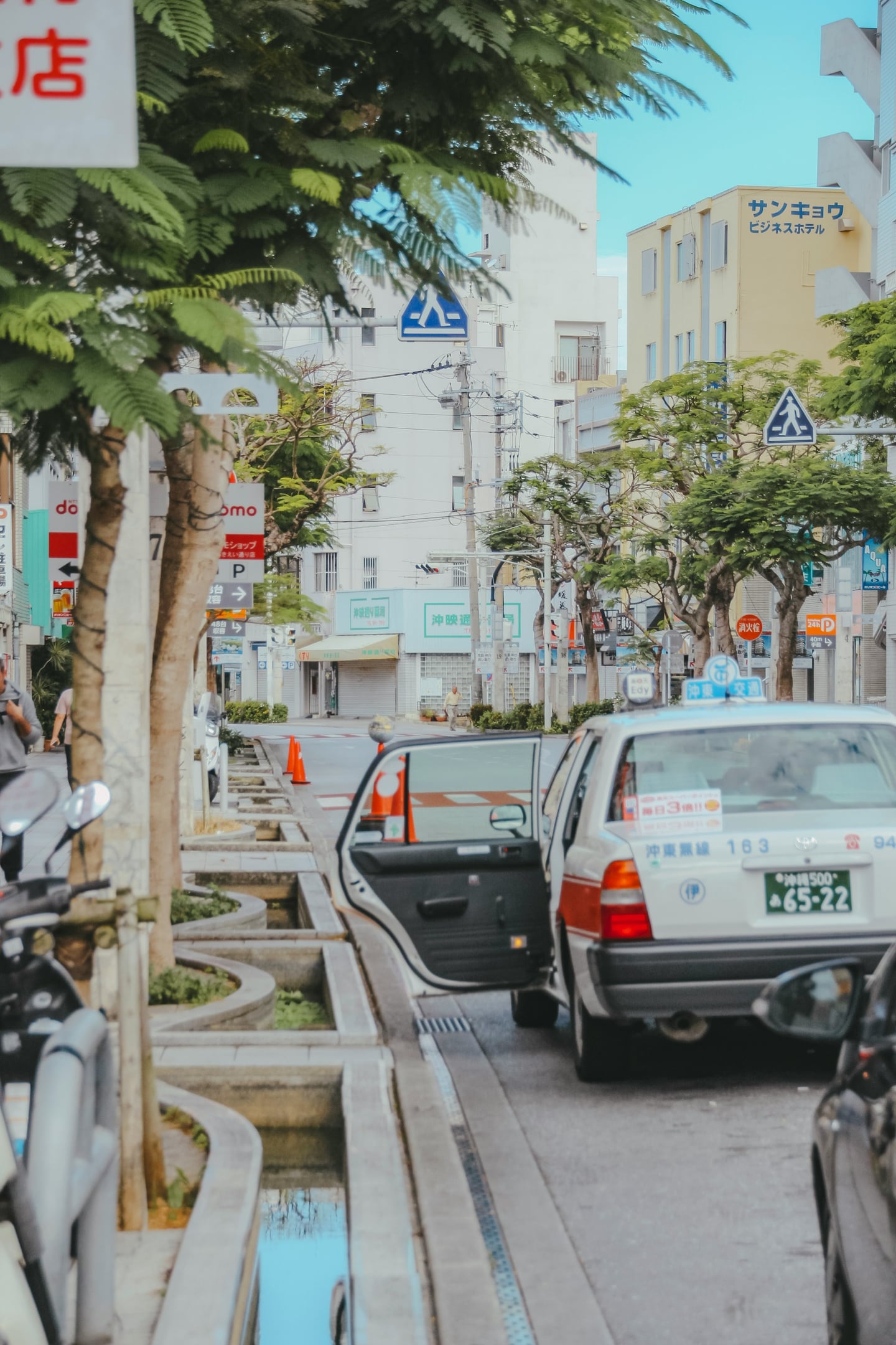 오키나와 국제거리, 푸른 가로수 사이로 문이 열린 택시와 일본어 간판이 늘어선 활기찬 나하 시내의 일상적인 풍경입니다.