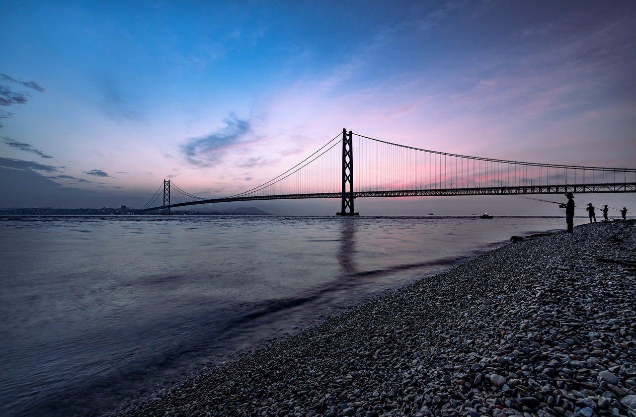 고베 아카시 해협 대교, 붉게 물든 노을을 배경으로 자갈 해변에서 낚시하는 사람들과 거대한 현수교의 실루엣이 조화를 이룹니다.