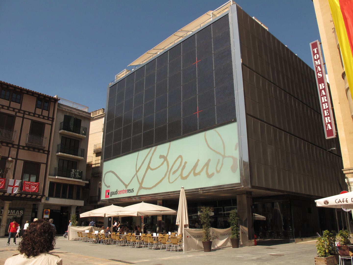 레우스 가우디 센터, 푸른 하늘 아래 가우디의 서명이 선명하게 새겨진 현대적인 박물관 외관과 활기찬 야외 테라스 카페의 풍경입니다.