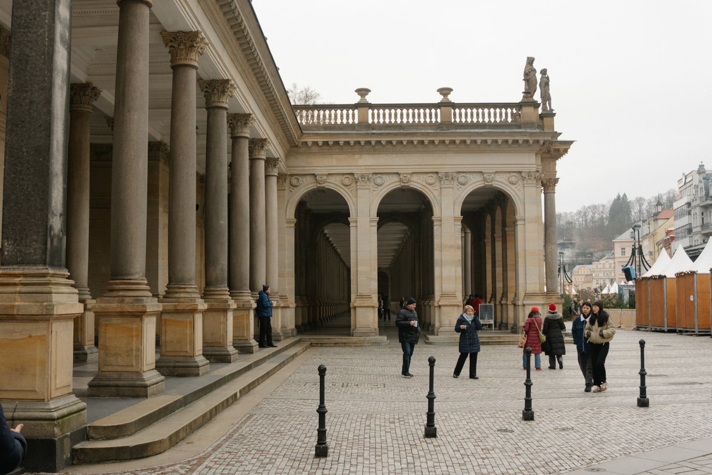 카를로비바리 믈린스카 콜로나다, 고전적인 석조 기둥과 아치형 복도가 길게 이어지는 웅장한 네오르네상스 건축물 앞에서 산책하는 사람들의 모습입니다.