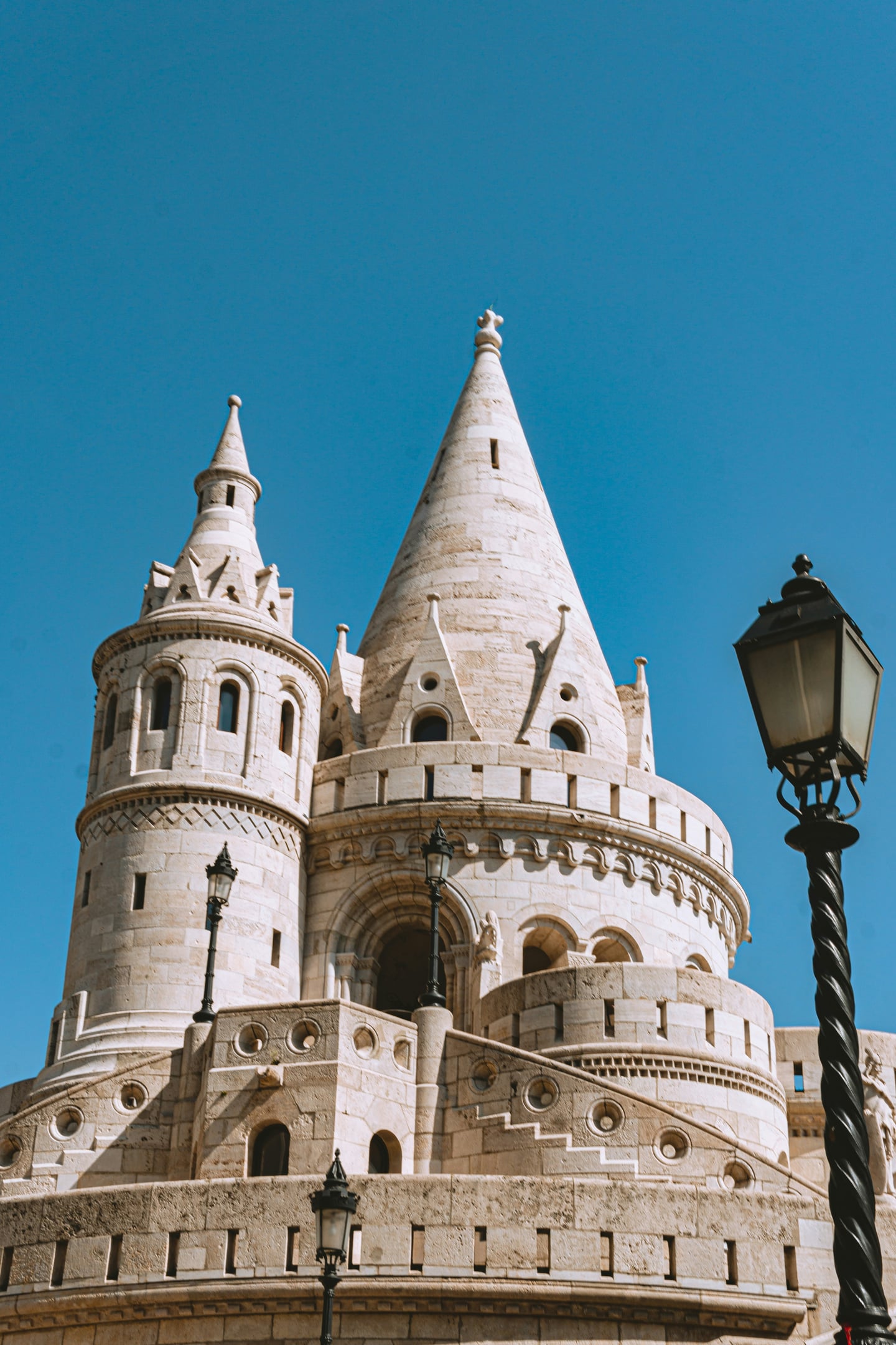 부다페스트 어부의 요새, 맑은 하늘 아래 우뚝 솟은 하얀 석조 탑과 고전적인 가로등이 어우러진 이국적인 풍경입니다.
