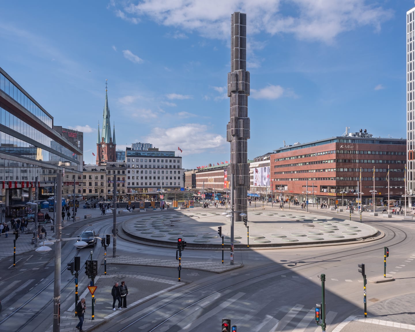 스톡홀름 세르옐스 광장, 푸른 하늘 아래 우뚝 솟은 거대한 유리 오벨리스크와 현대적인 도심 건물이 어우러진 탁 트인 전경입니다.