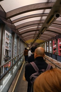 홍콩 미드레벨 에스컬레이터, 투명한 아치형 지붕 아래로 길게 뻗은 에스컬레이터를 타고 도심 사이를 이동하는 사람들의 일상적인 풍경입니다.