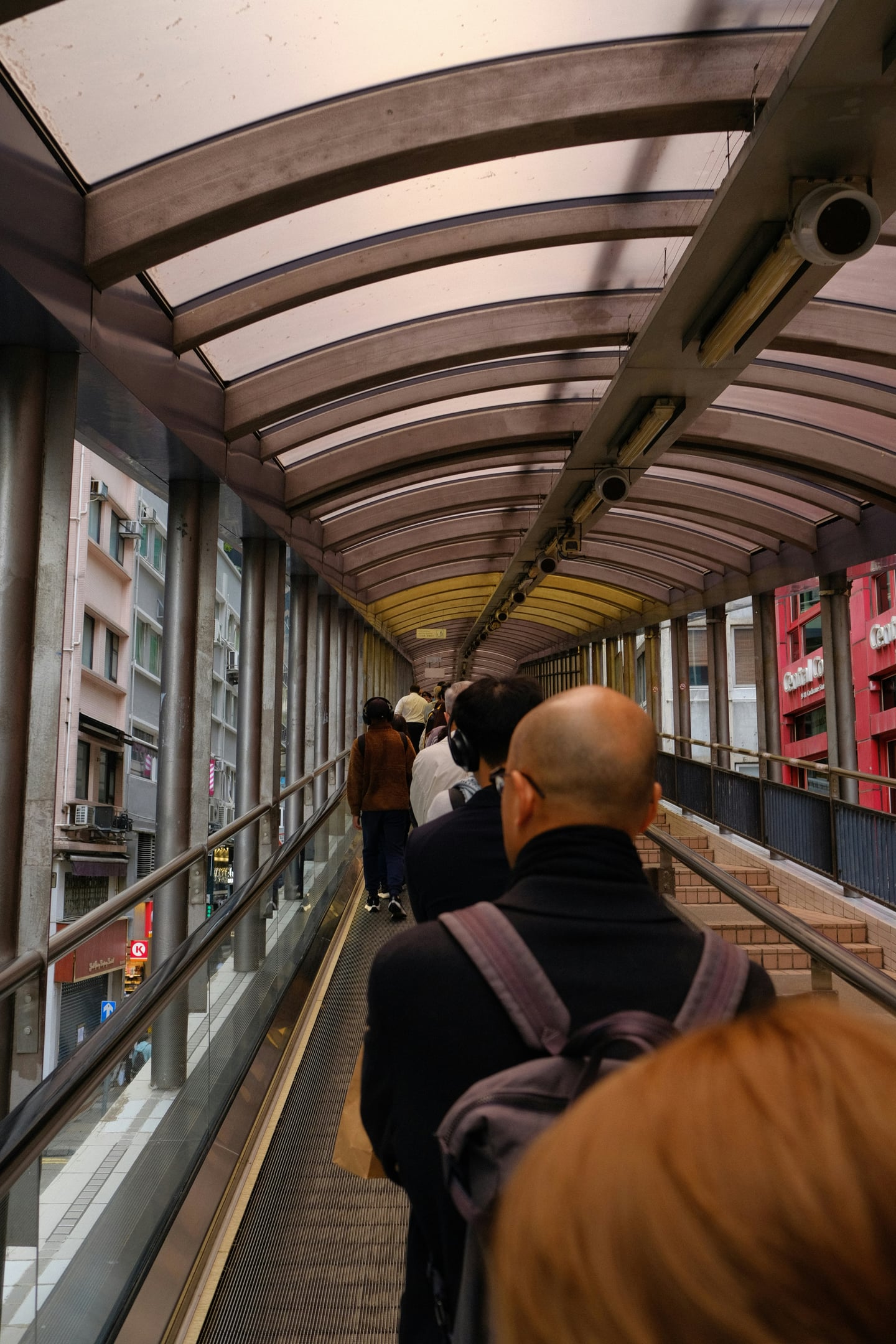 홍콩 미드레벨 에스컬레이터, 투명한 아치형 지붕 아래로 길게 뻗은 에스컬레이터를 타고 도심 사이를 이동하는 사람들의 일상적인 풍경입니다.