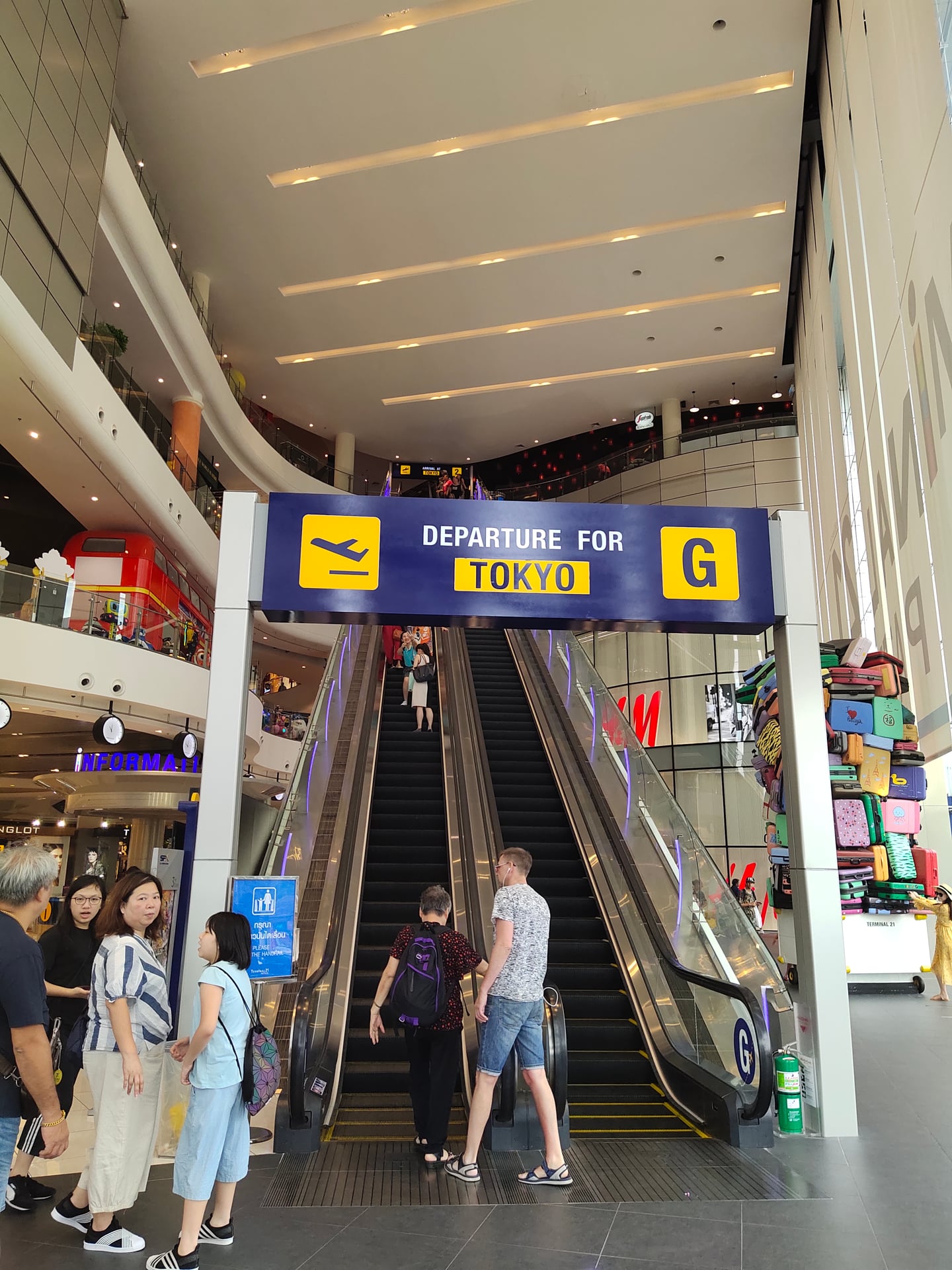파타야 터미널 21, 공항 게이트를 연상시키는 도쿄행 안내판과 에스컬레이터를 이용하는 방문객들의 활기찬 쇼핑몰 내부 전경입니다.