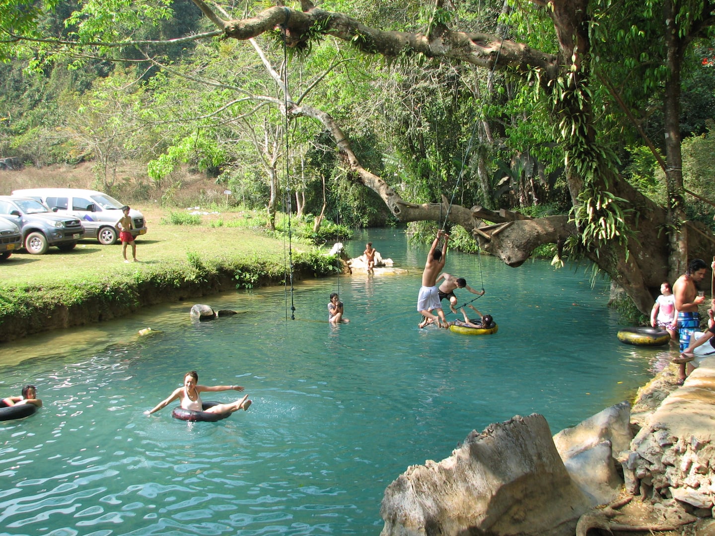 방비엥 블루라군, 울창한 나무 아래 에메랄드빛 맑은 물에서 튜브를 타고 밧줄 다이빙을 즐기며 휴식을 취하는 여행객들의 활기찬 모습입니다.