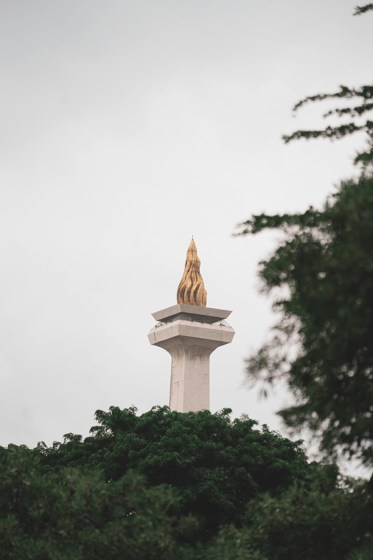 자카르타 모나스 광장, 울창한 초록빛 나무들 사이로 우뚝 솟은 국립기념탑의 황금빛 불꽃 조형물이 흐린 하늘 아래 웅장하게 자리 잡고 있습니다.