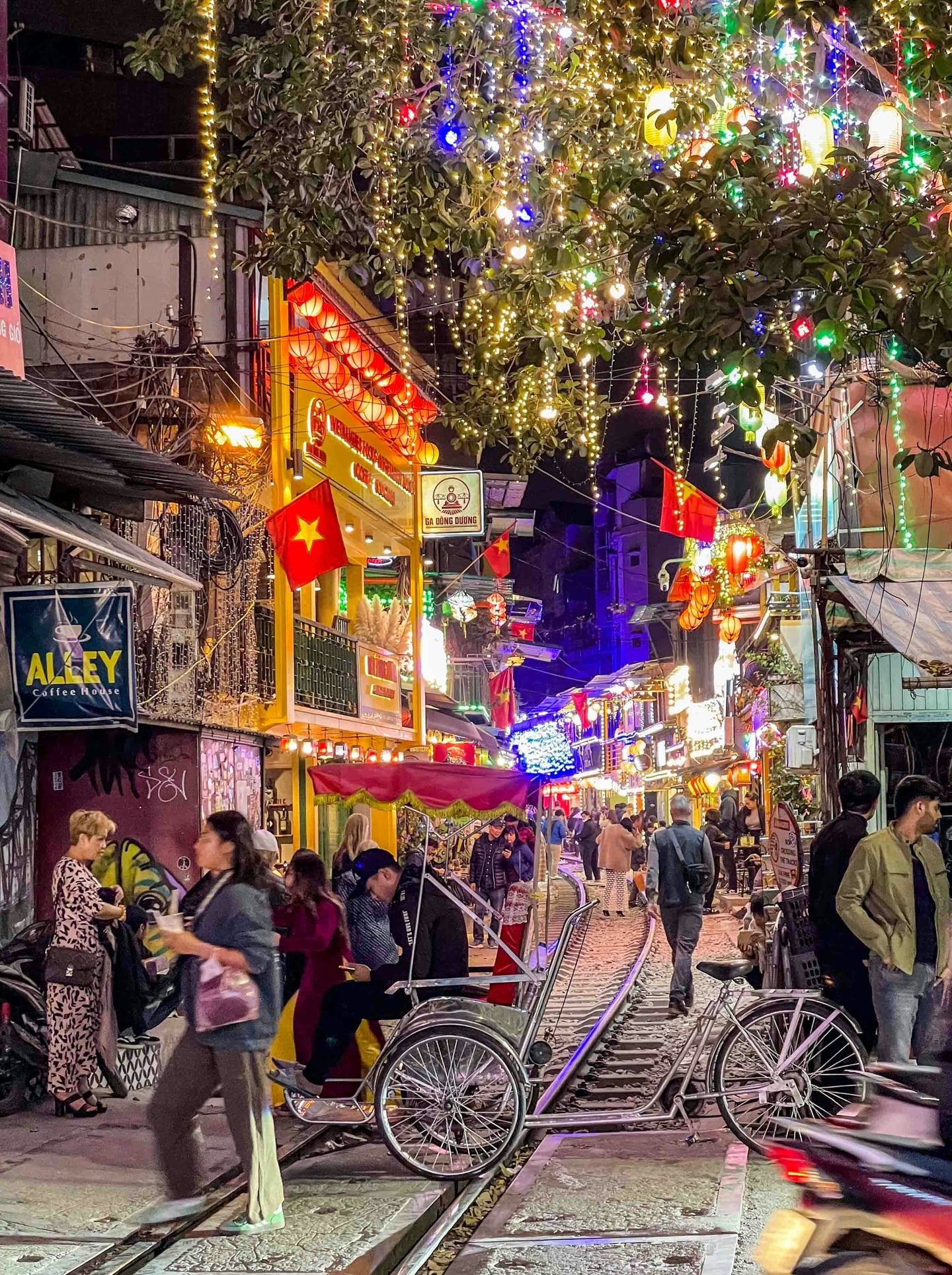 하노이 하노이 여행자 거리, 화려한 전구 조명과 붉은 베트남 국기가 가득한 기찻길 옆 카페 거리에서 밤을 즐기는 관광객들의 활기찬 모습.