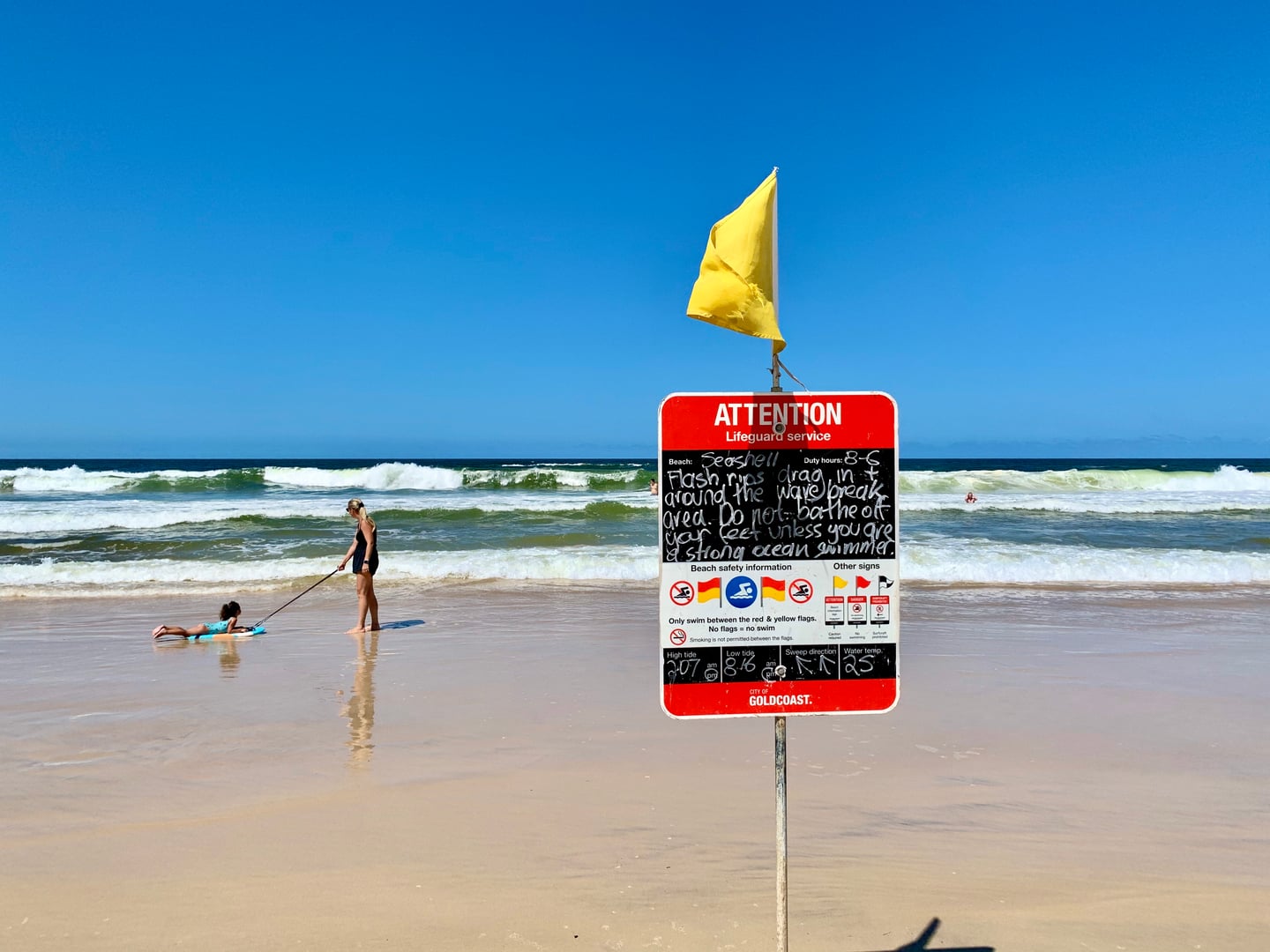 골드코스트 머메이드 비치, 노란색 안전 깃발과 표지판 너머로 파도를 즐기는 사람들과 푸른 바다 풍경이 펼쳐집니다.