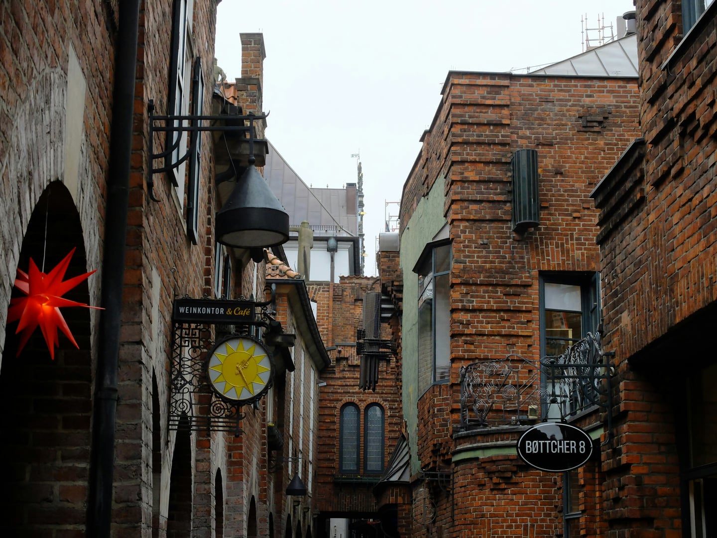 브레멘 뵈트허 거리, 붉은 벽돌 건물들 사이로 보이는 고풍스러운 철제 간판과 독특한 해 모양의 장식이 어우러진 중세풍 골목의 풍경입니다.