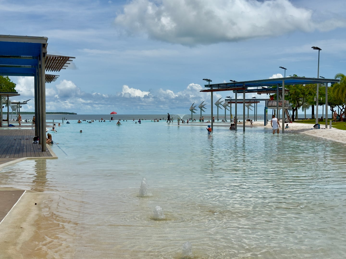 케언즈 케언즈 라군, 푸른 하늘 아래 넓게 펼쳐진 야외 수영장에서 여유롭게 물놀이를 즐기는 사람들과 평화로운 해변 풍경.