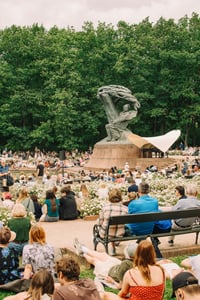 바르샤바 쇼팽 공원, 울창한 숲과 화사한 장미꽃 사이에서 쇼팽 동상을 배경으로 야외 음악회를 즐기는 사람들의 평화로운 모습입니다.
