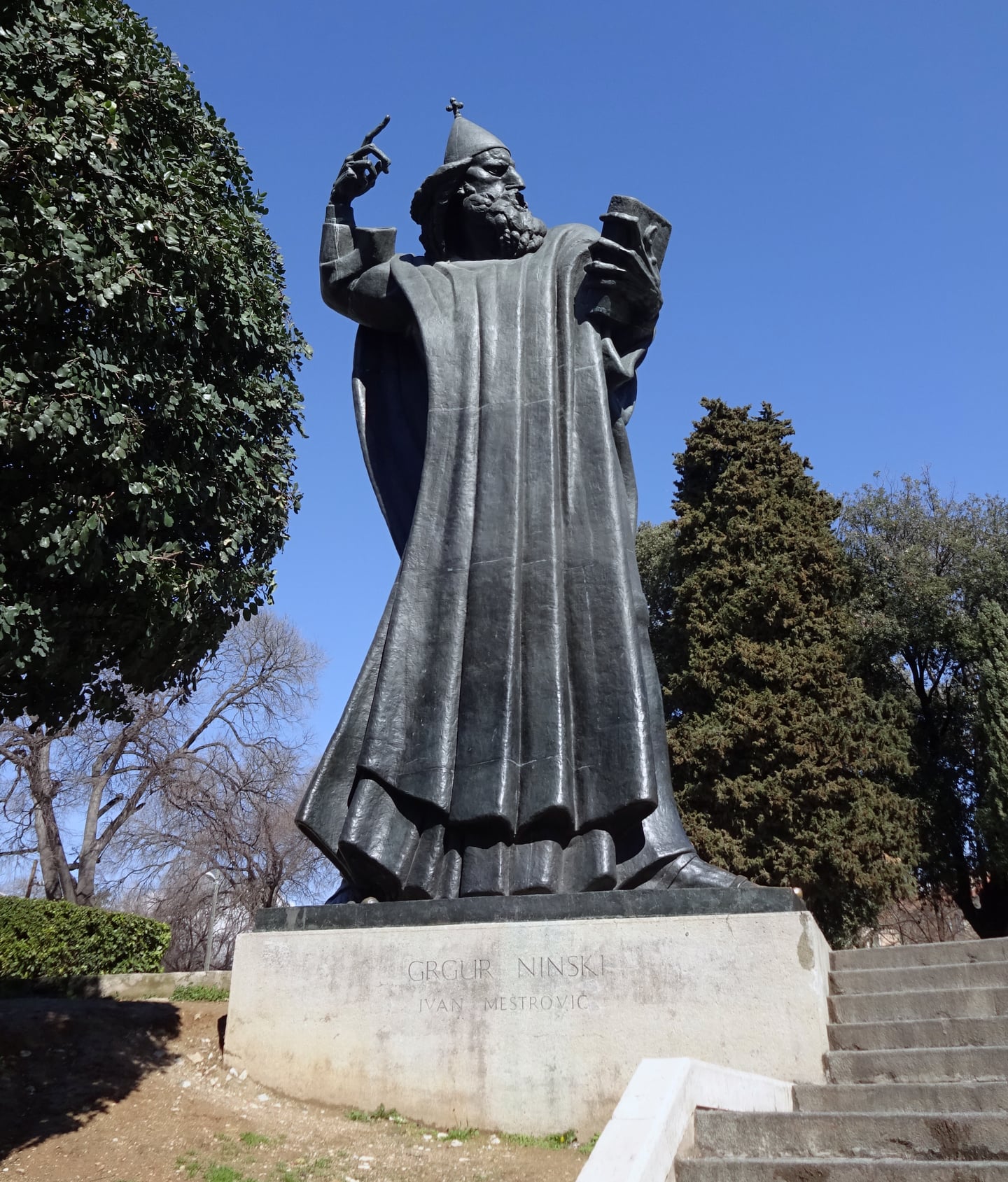 스플리트 그레고리우스 닌 동상, 푸른 하늘 아래 위엄 있게 서 있는 거대한 청동상의 전신과 정교한 조각 디테일이 돋보이는 풍경입니다.