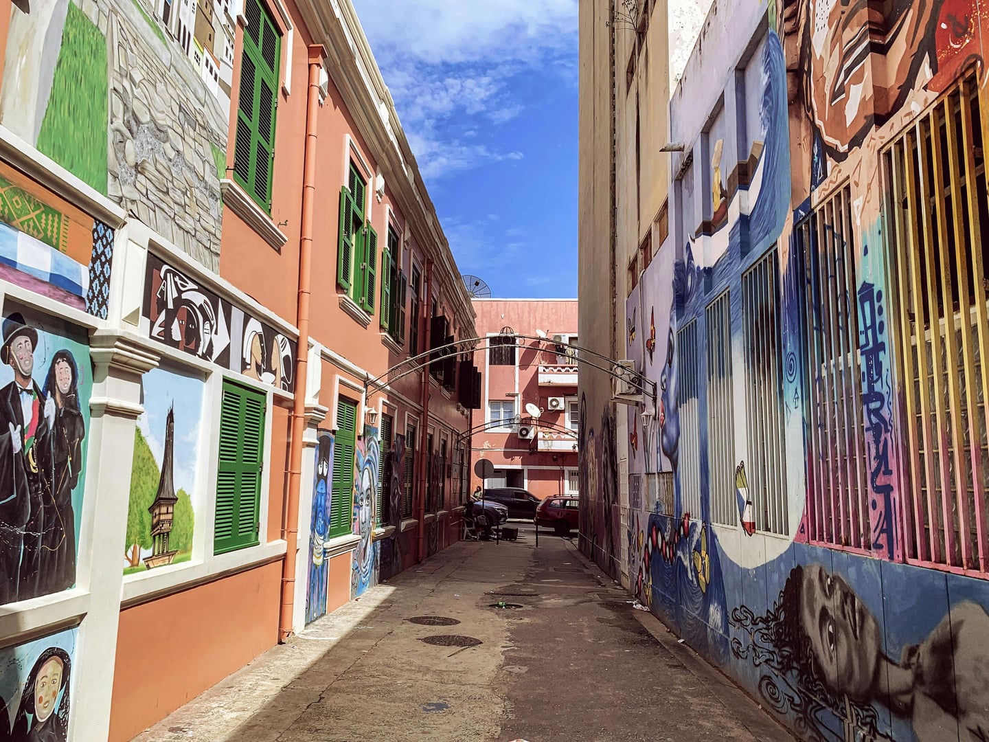 범용 골목 벽화 여성, 화려한 색채의 인물 벽화와 그라피티가 가득한 좁은 골목길 사이로 푸른 하늘이 펼쳐진 이국적인 풍경입니다.