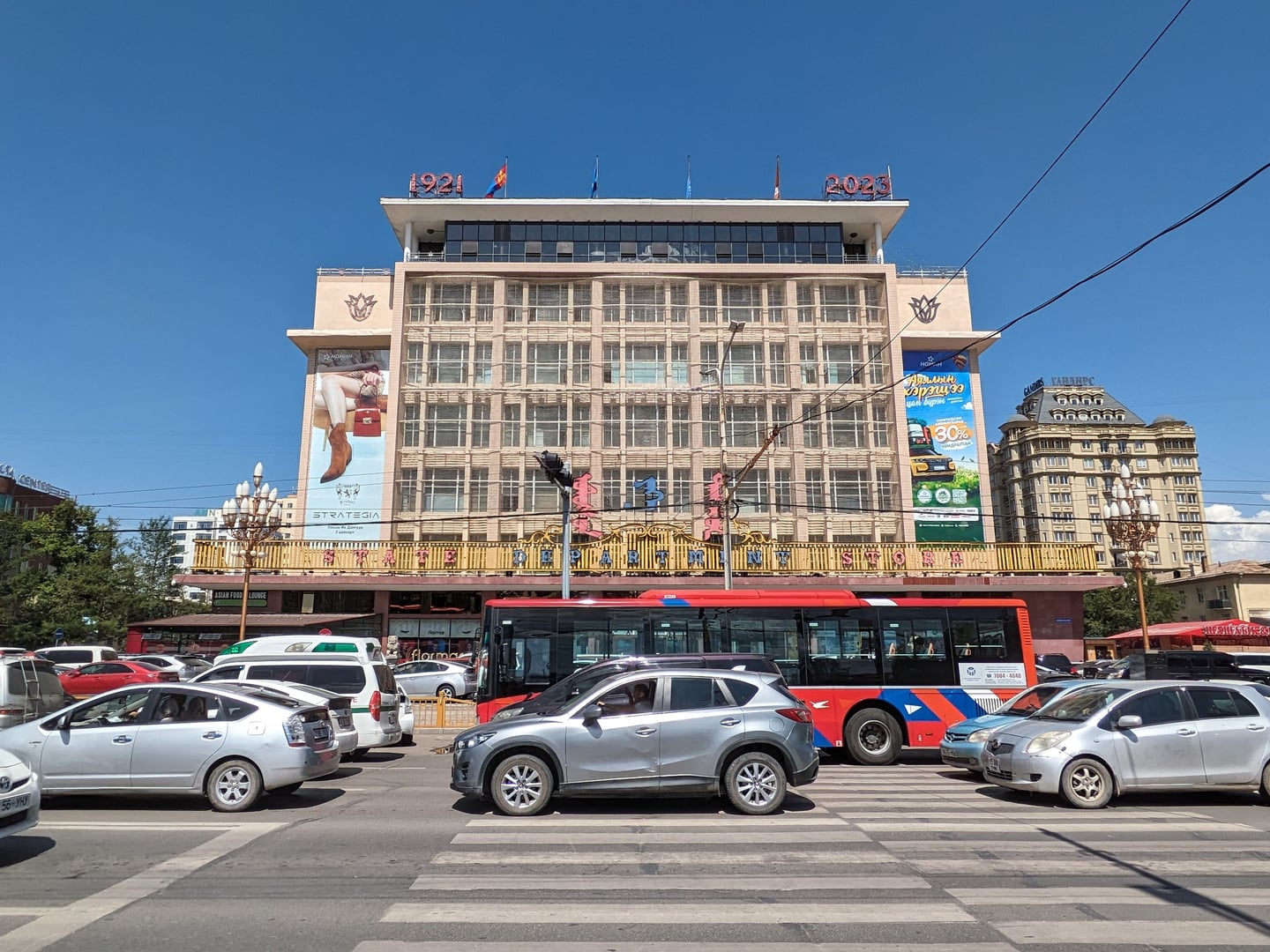 울란바토르 국영 백화점, 맑은 하늘 아래 고전적인 외관의 백화점 건물과 도로 위를 지나는 붉은색 버스가 어우러진 도심 풍경입니다.