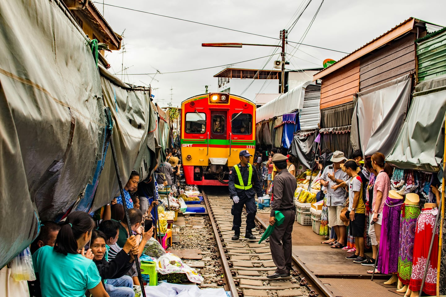방콕 매끌렁 기찻길 시장, 좁은 철로 위로 노란색과 빨간색 기차가 상점 천막 사이를 아슬아슬하게 통과하는 이색적인 풍경입니다.