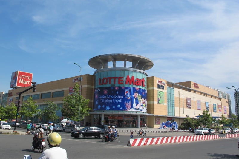 호치민 롯데마트, 맑은 하늘 아래 대형 로고와 광고판이 돋보이는 현대적인 쇼핑몰 외관과 활기찬 도로 풍경입니다.
