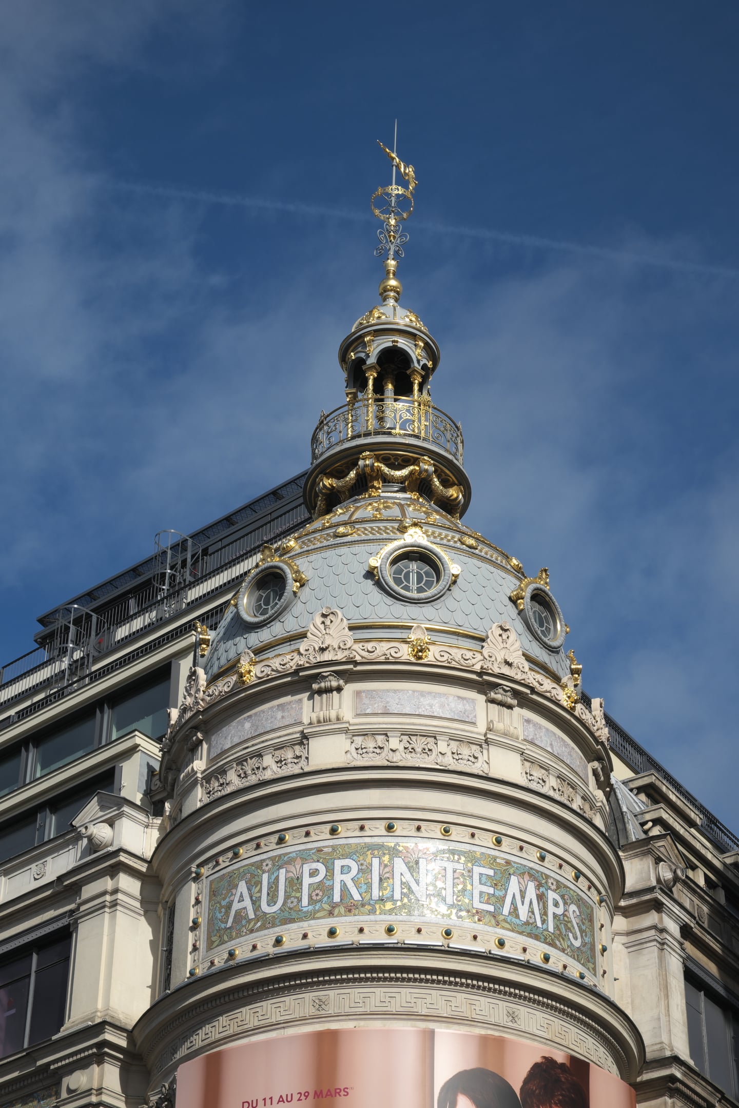 파리 쁘렝땅 백화점, 푸른 하늘 아래 금빛 장식으로 빛나는 화려한 아르누보 양식의 돔 지붕과 고풍스러운 외벽 문구.