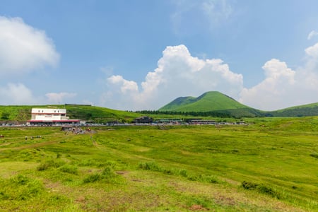 아소 아소산, 푸른 초원이 끝없이 펼쳐진 쿠사센리 평원 너머로 웅장한 산맥과 화산 박물관이 어우러진 맑은 여름날의 풍경입니다.