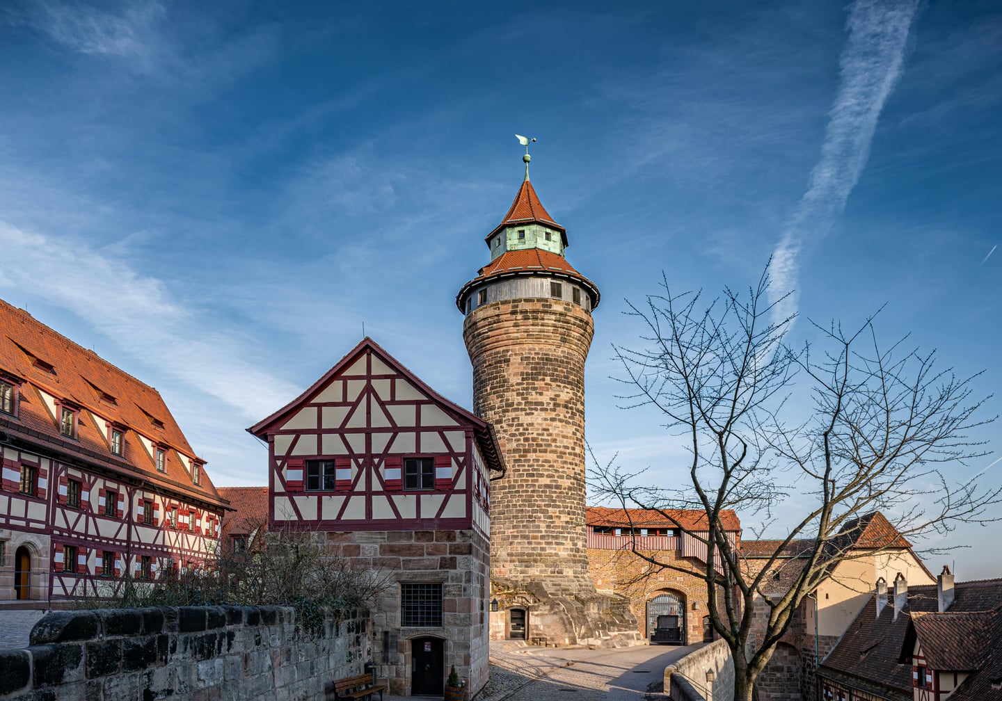 뉘른베르크 카이저부르크, 푸른 하늘 아래 우뚝 솟은 원형의 진벨 탑과 붉은 지붕의 전통 목조 가옥이 어우러진 중세 성채의 전경입니다.