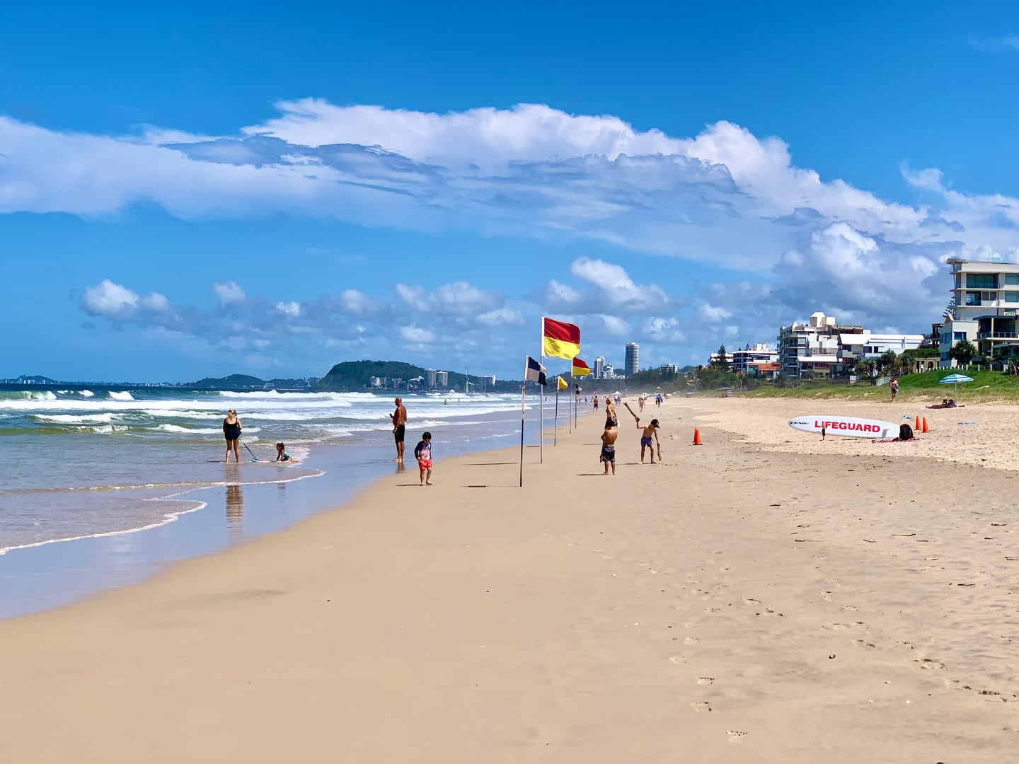 골드코스트 머메이드 비치, 맑은 하늘 아래 라이프가드 깃발이 펄럭이는 황금빛 모래사장과 여유롭게 해수욕을 즐기는 사람들의 모습입니다.