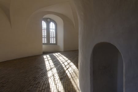 코펜하겐 원형탑, 하얀 곡선 벽면과 벽돌 바닥 위로 창가 햇살이 격자무늬 그림자를 드리우는 나선형 경사로의 고요한 풍경입니다.