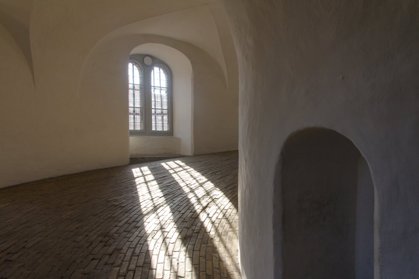 코펜하겐 원형탑, 하얀 곡선 벽면과 벽돌 바닥 위로 창가 햇살이 격자무늬 그림자를 드리우는 나선형 경사로의 고요한 풍경입니다.