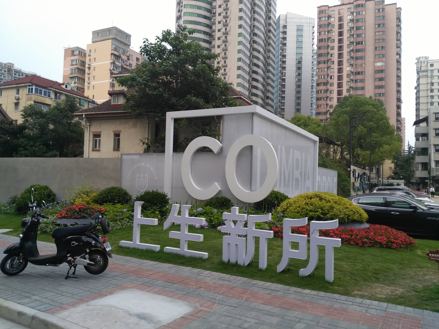 상하이 콜롬비아 서클, 현대적인 고층 빌딩 숲 사이로 세련된 흰색 로고 조형물과 화사한 꽃들이 어우러진 입구 광장의 모습입니다.