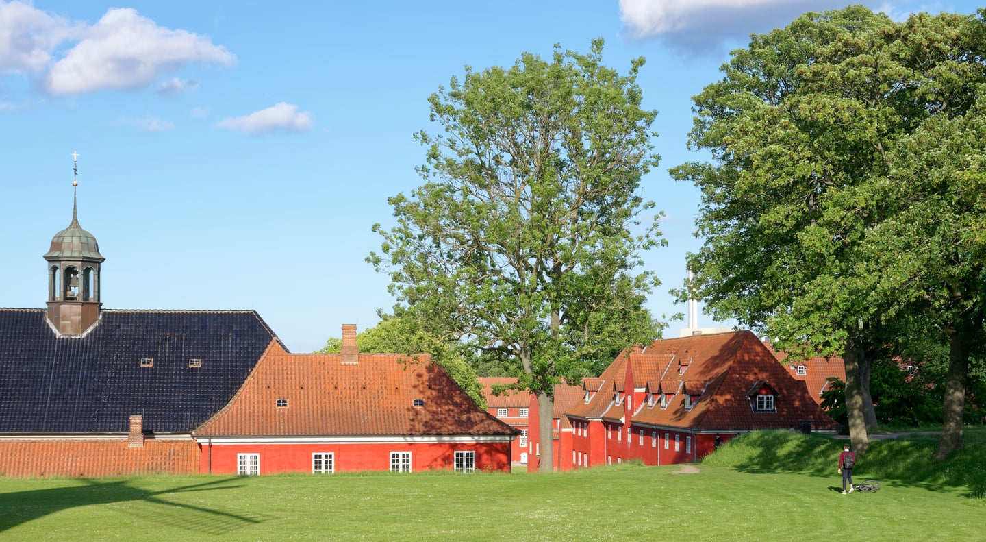 코펜하겐 카스텔레트 요새, 푸른 잔디 언덕 너머로 보이는 붉은색 전통 가옥들과 평화로운 북유럽의 풍경이 인상적입니다.