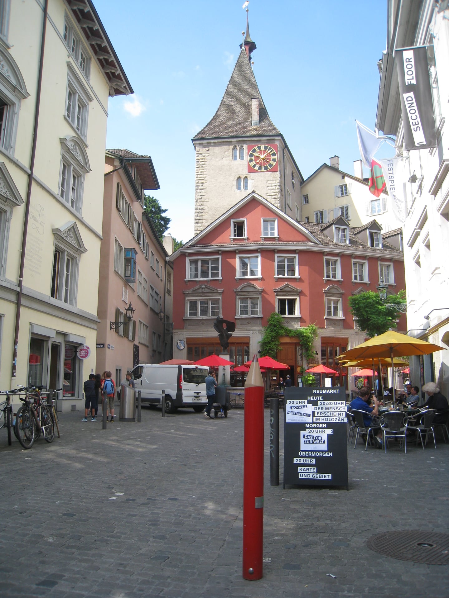 취리히 니더도르프 거리, 고풍스러운 붉은 건물과 시계탑을 배경으로 노천 카페와 빨간 연필 조형물이 어우러진 구시가지 풍경입니다.