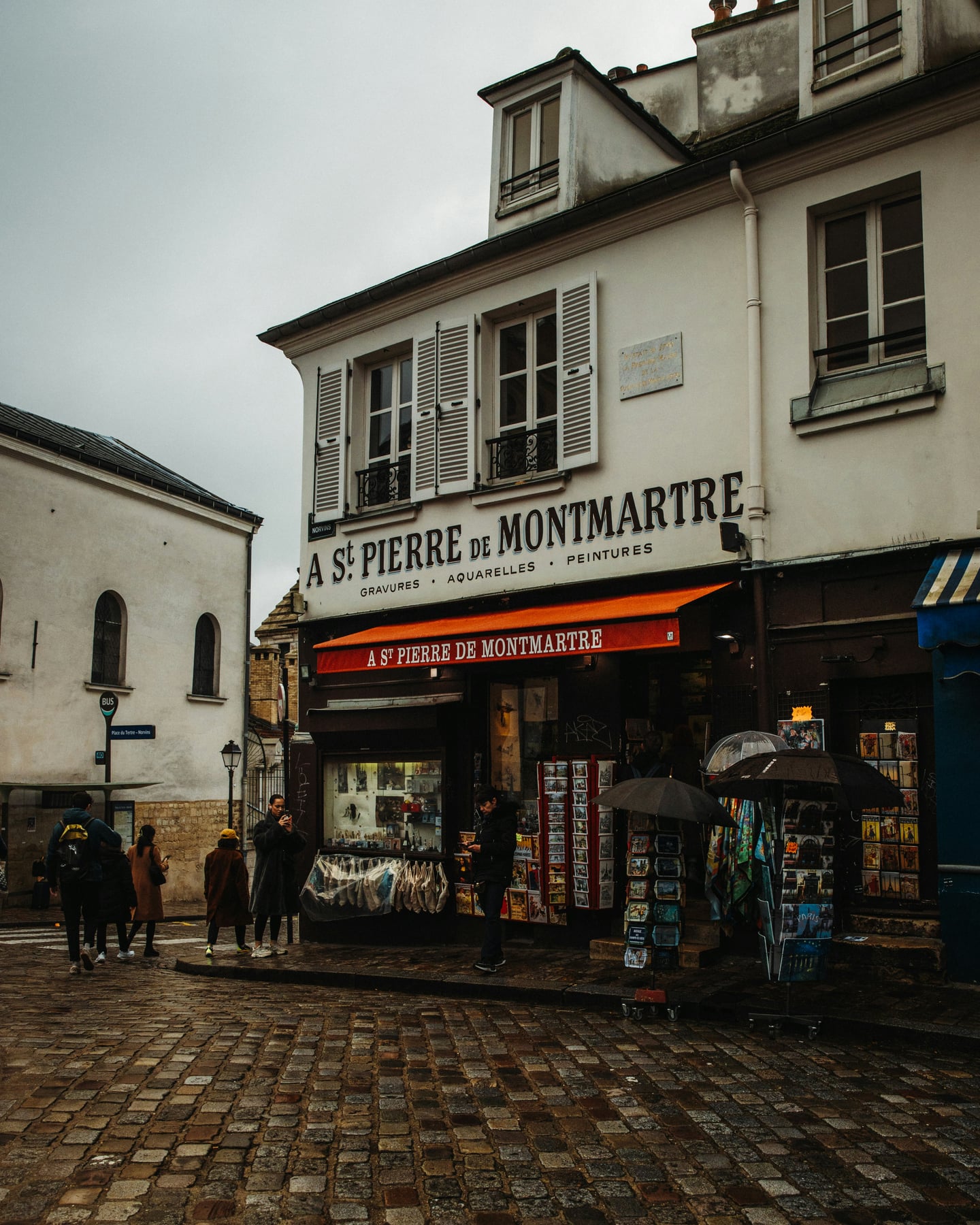 파리 테르트르 광장, 비에 젖은 고풍스러운 돌바닥 거리와 붉은 차양이 돋보이는 몽마르트르 기념품점의 서정적인 풍경입니다.