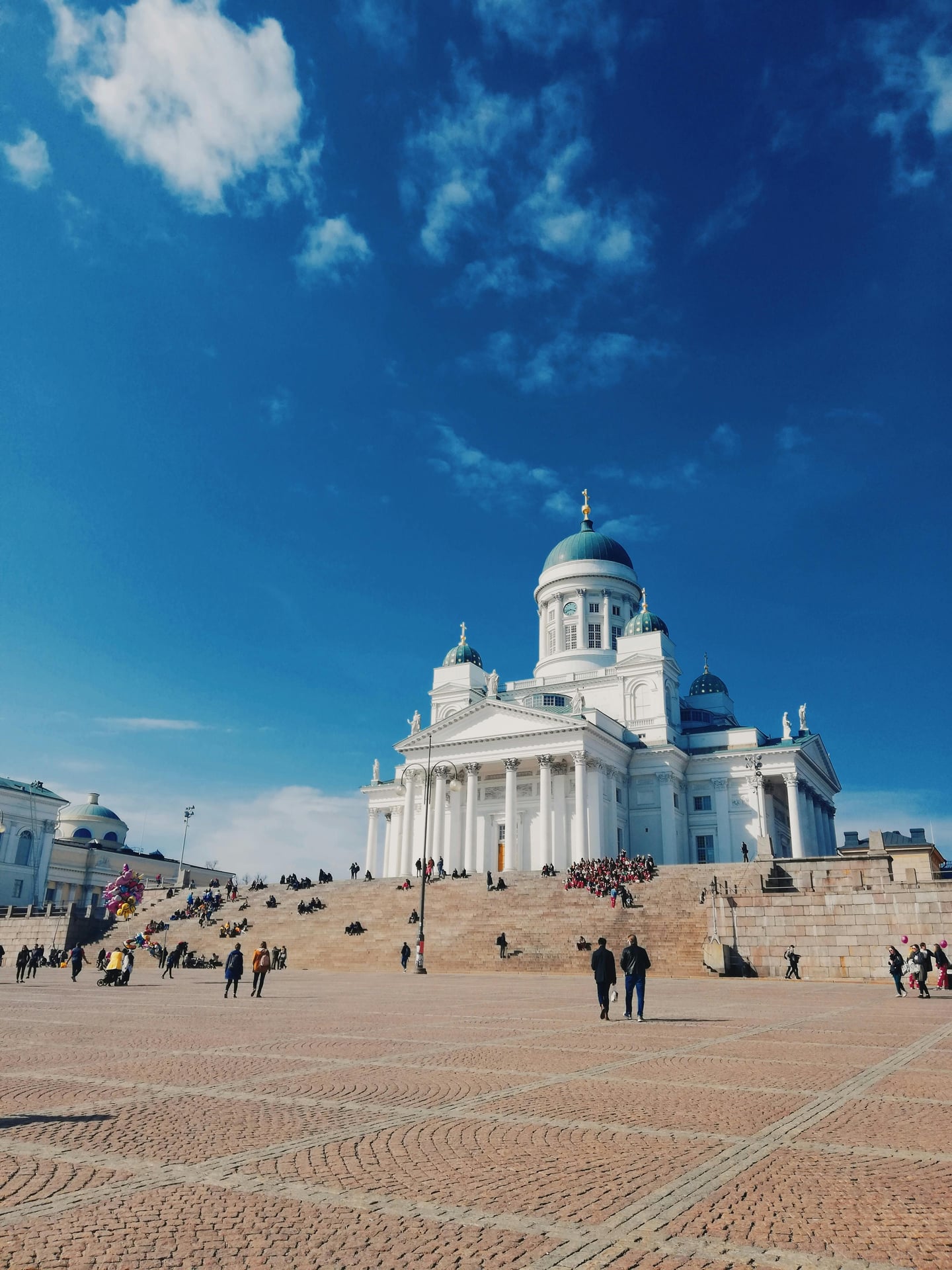 헬싱키 원로원 광장, 푸른 하늘 아래 웅장하게 솟은 하얀 대성당과 계단에 모여 휴식을 취하는 사람들의 평화로운 풍경입니다.