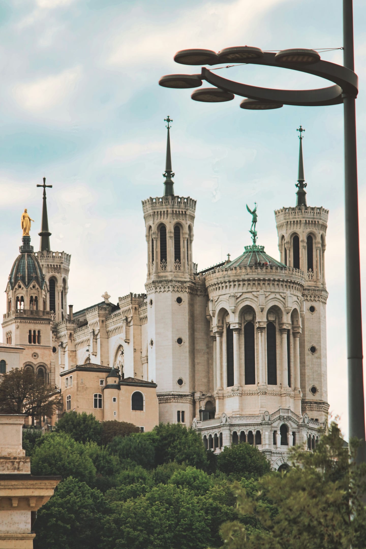 리옹 푸르비에르 대성당, 푸른 숲 위로 솟아오른 웅장한 흰색 첨탑과 화려한 금빛 성모 마리아 상이 어우러진 고풍스러운 풍경입니다.