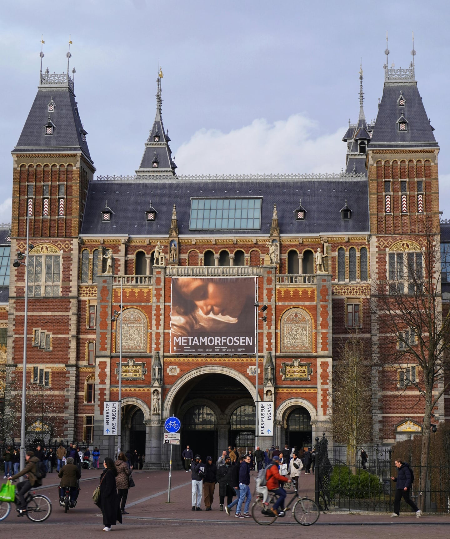 암스테르담 국립미술관, 웅장한 붉은 벽돌 건물 외관과 입구 주변에서 자전거를 타며 활기차게 움직이는 사람들의 풍경입니다.