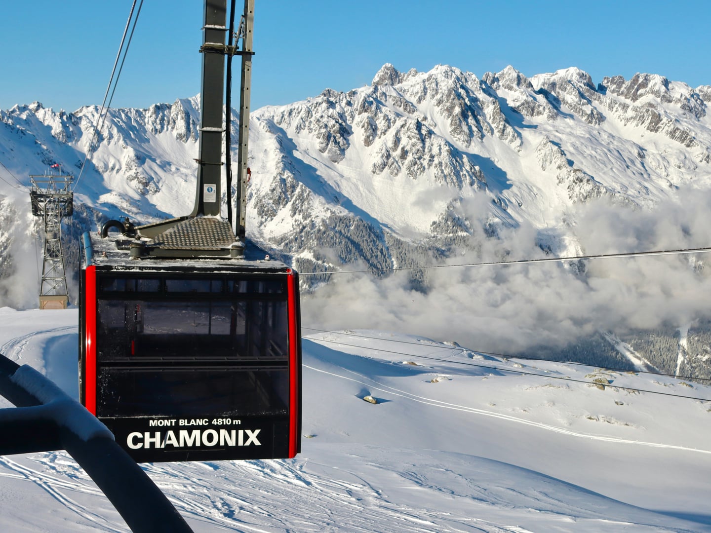 샤모니 몽블랑, 눈 덮인 알프스 산맥을 배경으로 CHAMONIX 로고가 새겨진 빨간색 케이블카가 설산 위를 가로지르는 풍경입니다.