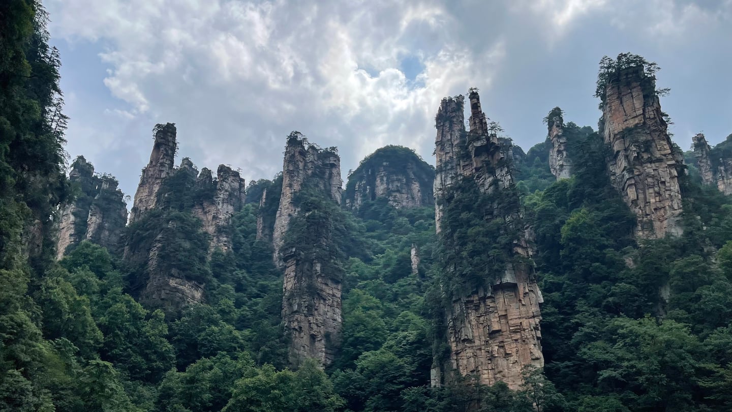 장가계 무릉원 풍경구, 울창한 숲 위로 솟아오른 거대한 수직 바위 기둥들과 구름 낀 하늘이 어우러진 웅장한 산세.