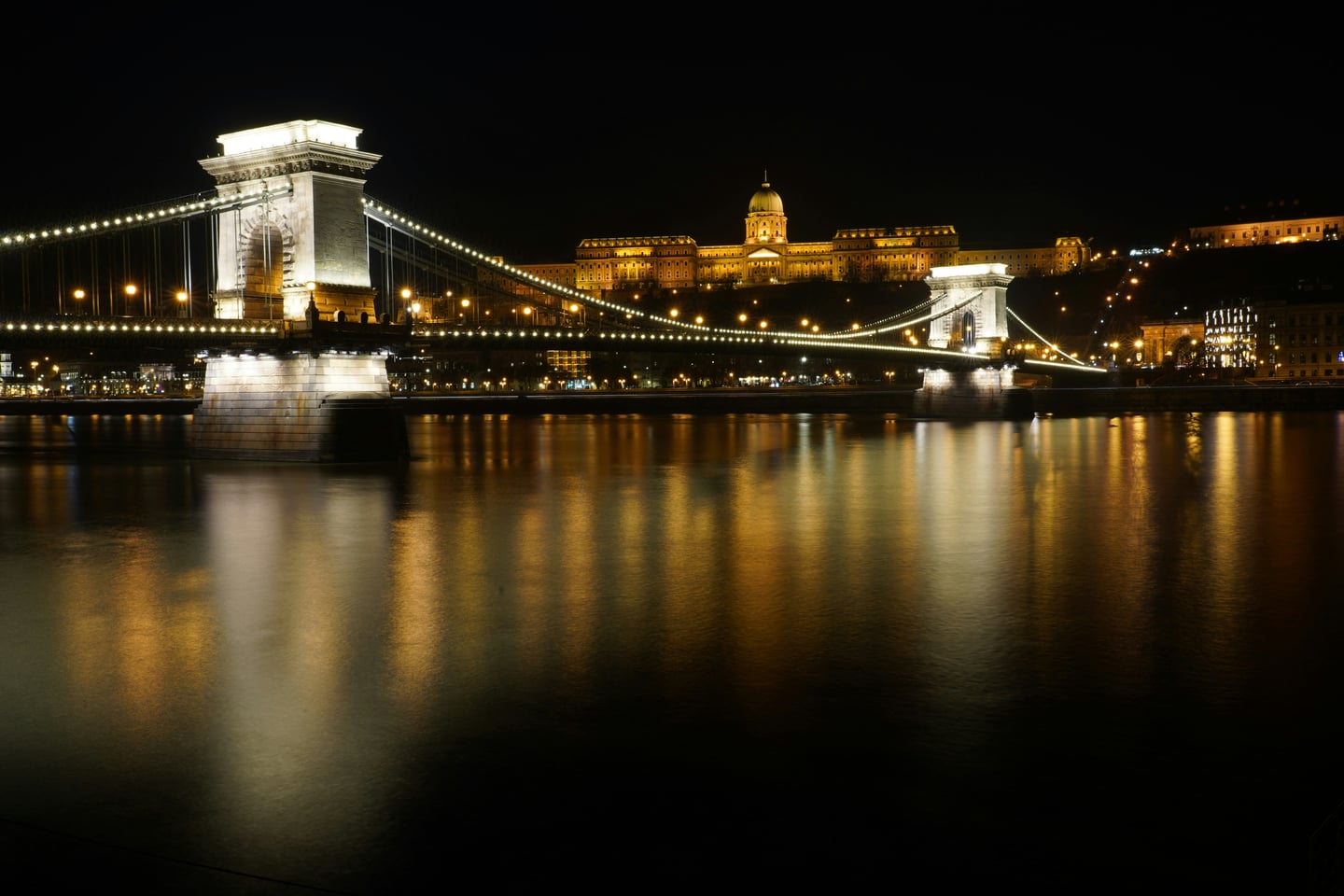부다페스트 세체니 다리, 어두운 밤 다뉴브 강물 위로 황금빛 조명이 화려하게 빛나는 현수교와 부다 성의 야경입니다.