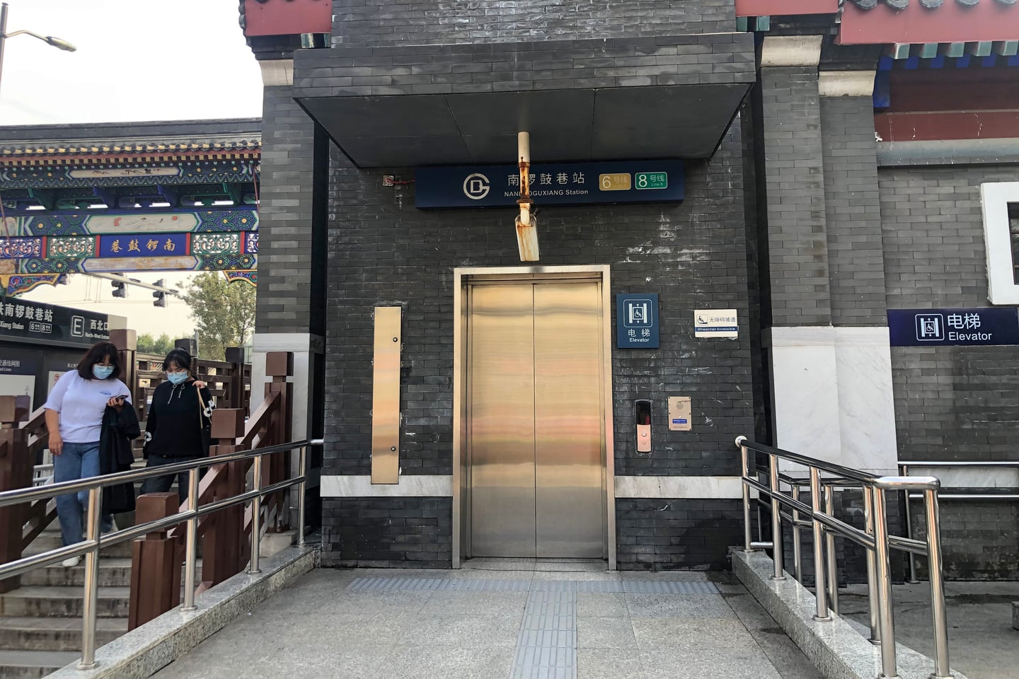 베이징 난뤄구샹, 전통적인 회색 벽돌 건물에 설치된 현대적인 지하철역 엘리베이터 입구와 주변 행인들의 모습입니다.
