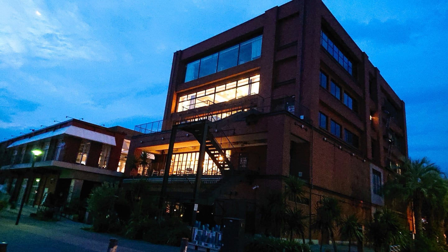 요코하마 마린 앤 워크, 해 질 녘 푸른 하늘 아래 붉은 벽돌 건물의 따뜻한 조명과 세련된 외관이 돋보이는 풍경입니다.