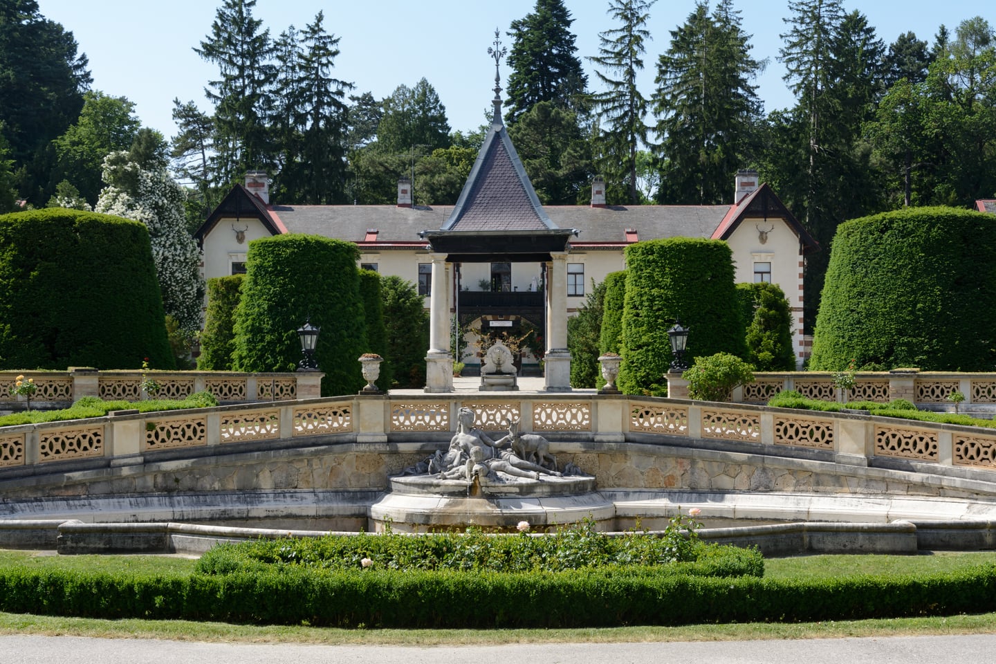 빈 헤르메스빌라, 푸른 숲을 배경으로 화려한 조각 분수와 대칭을 이루는 고전적인 건축물의 우아한 전경입니다.