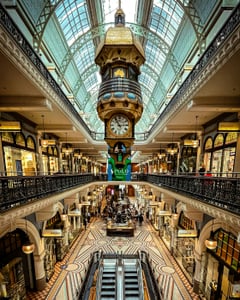 시드니 퀸 빅토리아 빌딩, 화려한 유리 돔 천장 아래 매달린 거대한 빈티지 시계와 고풍스러운 쇼핑몰 내부 전경입니다.