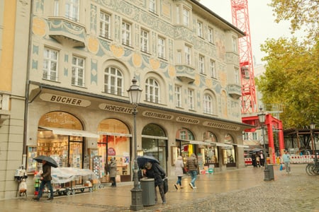 뮌헨 카우핑거 거리, 비 내리는 보행자 전용 도로 위로 화려한 벽화가 그려진 고풍스러운 건물과 활기찬 상점가 풍경이 펼쳐집니다.