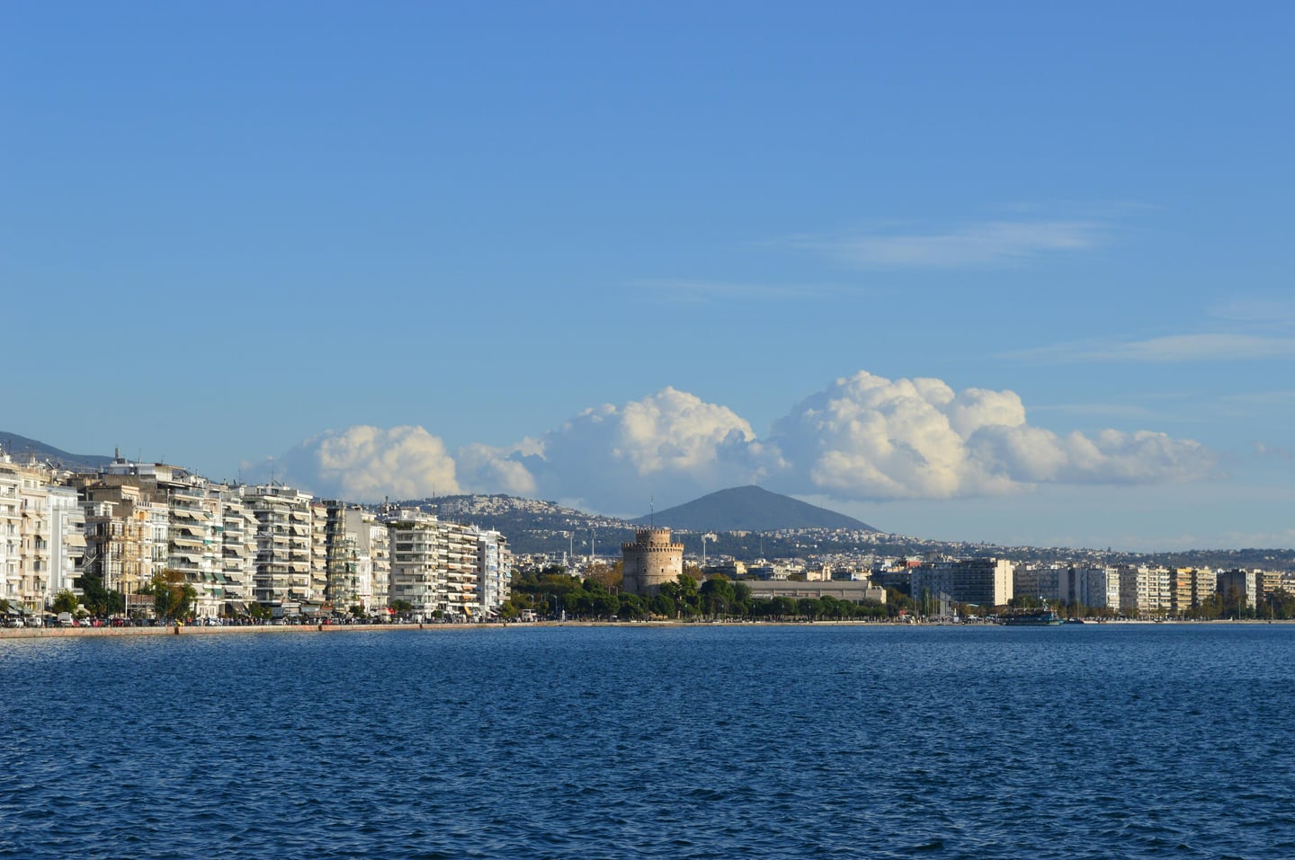 테살로니키 화이트 타워, 푸른 바다와 맞닿은 해안 산책로를 따라 우뚝 솟은 역사적인 원형 탑과 현대적인 도시 건물이 어우러진 풍경입니다.