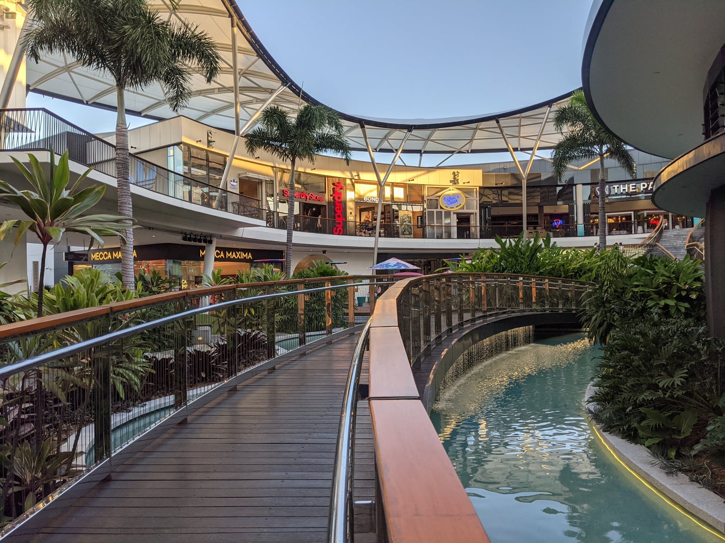 골드코스트 퍼시픽 페어, 인공 수로와 열대 식물이 어우러진 야외 쇼핑몰의 현대적인 목재 다리 풍경입니다.