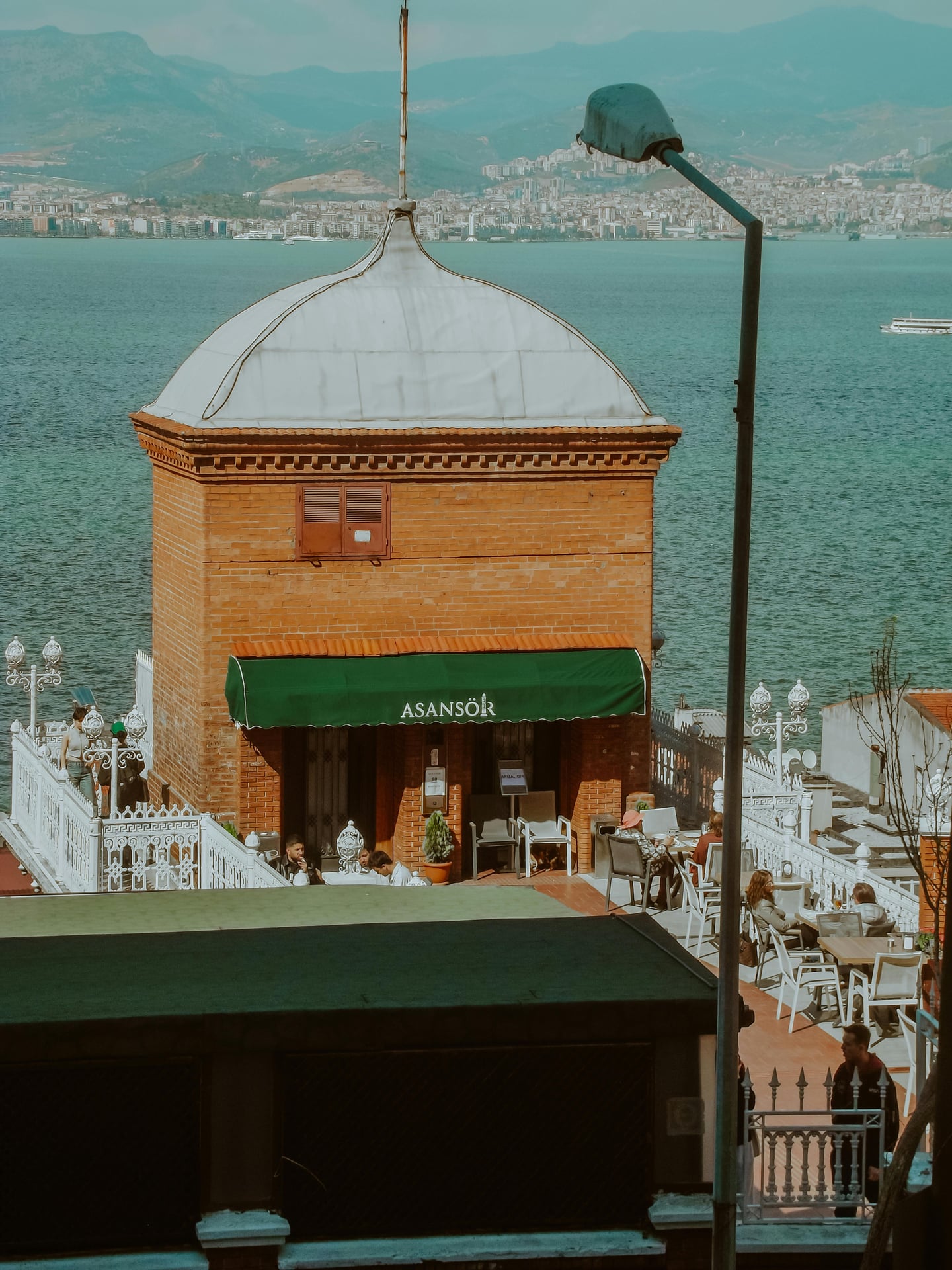 이즈미르 아산쇼르, 붉은 벽돌 타워와 푸른 바다가 어우러진 테라스 카페에서 사람들이 휴식을 취하는 풍경입니다.