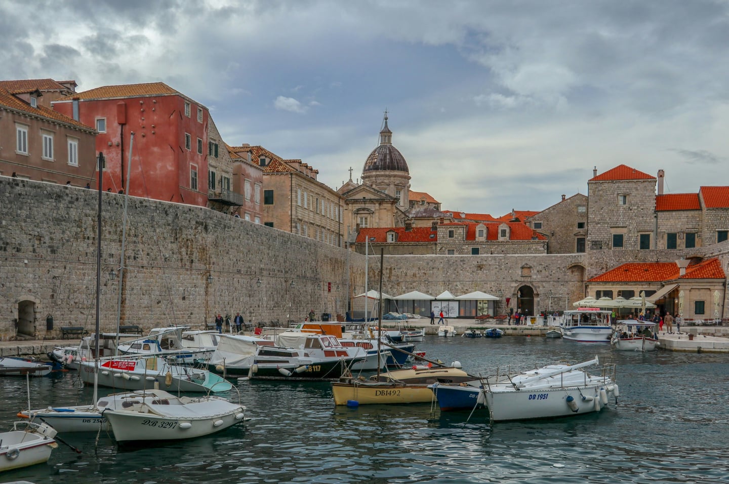 두브로브니크 구항구, 견고한 성벽 아래 정박한 작은 배들과 붉은 지붕의 고풍스러운 건물들이 어우러진 평화로운 항구의 전경입니다.