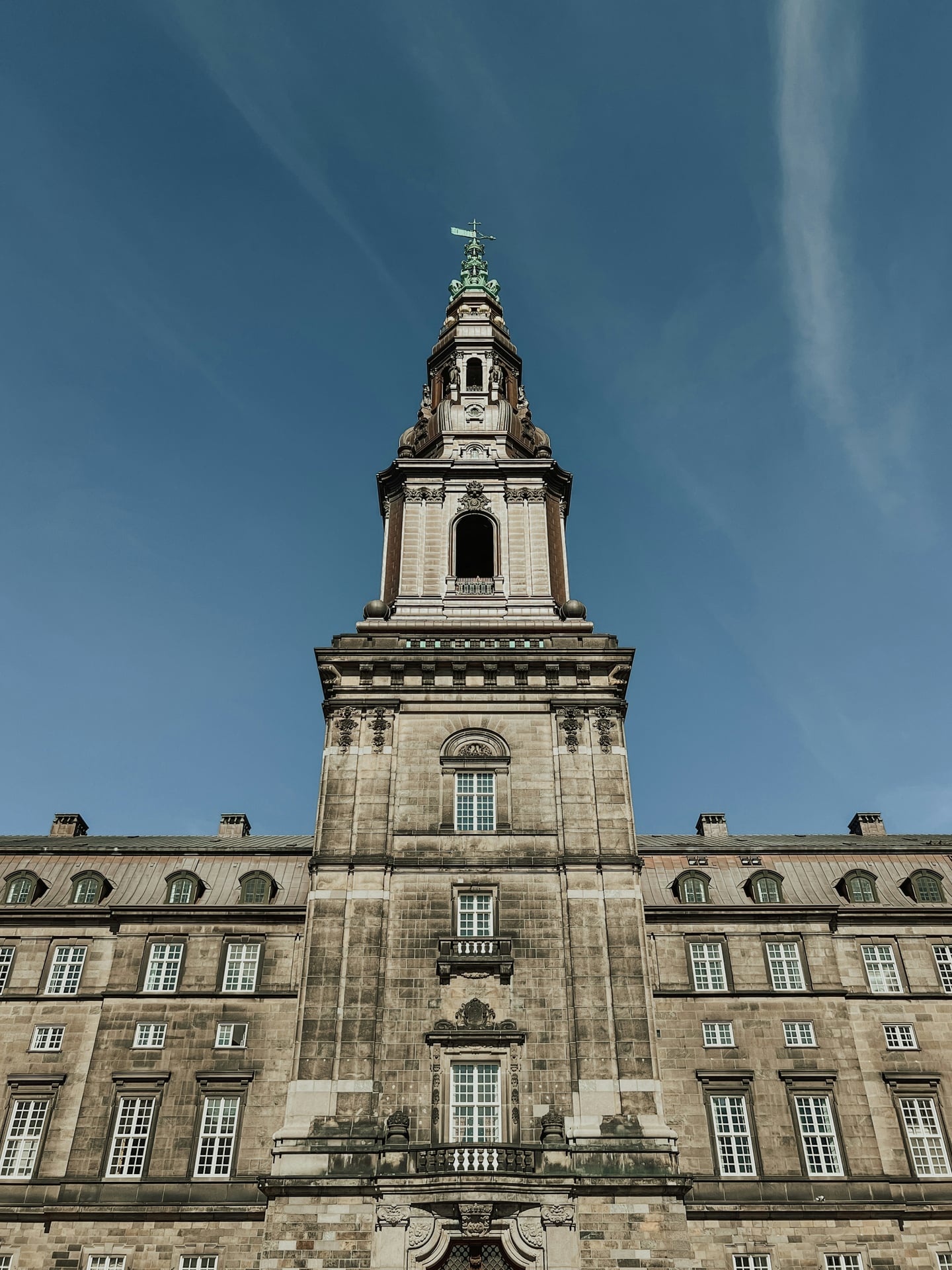 코펜하겐 크리스티안보르 궁전, 맑은 하늘 아래 높게 솟은 중앙 타워와 고풍스러운 석조 건물의 대칭미가 돋보이는 전경입니다.