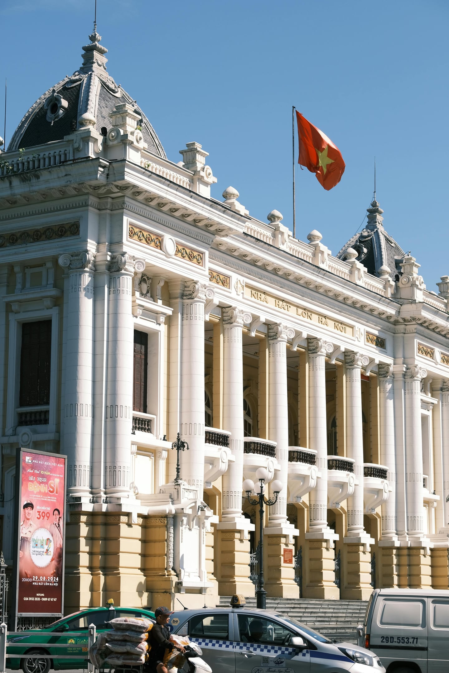 하노이 하노이 오페라 하우스, 푸른 하늘 아래 웅장한 신고전주의 양식의 기둥과 붉은 베트남 국기가 돋보이는 외관 전경입니다.