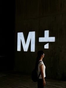 홍콩 M+, 어두운 콘크리트 벽면 위에서 밝게 빛나는 대형 로고 사인과 그 앞에 서 있는 관람객의 실루엣이 현대적인 분위기를 자아냅니다.
