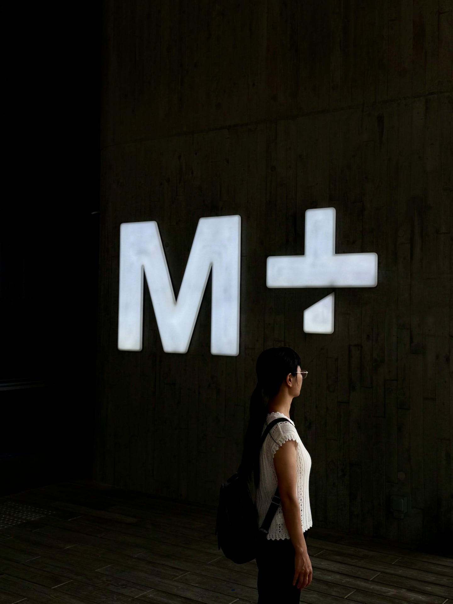 홍콩 M+, 어두운 콘크리트 벽면 위에서 밝게 빛나는 대형 로고 사인과 그 앞에 서 있는 관람객의 실루엣이 현대적인 분위기를 자아냅니다.